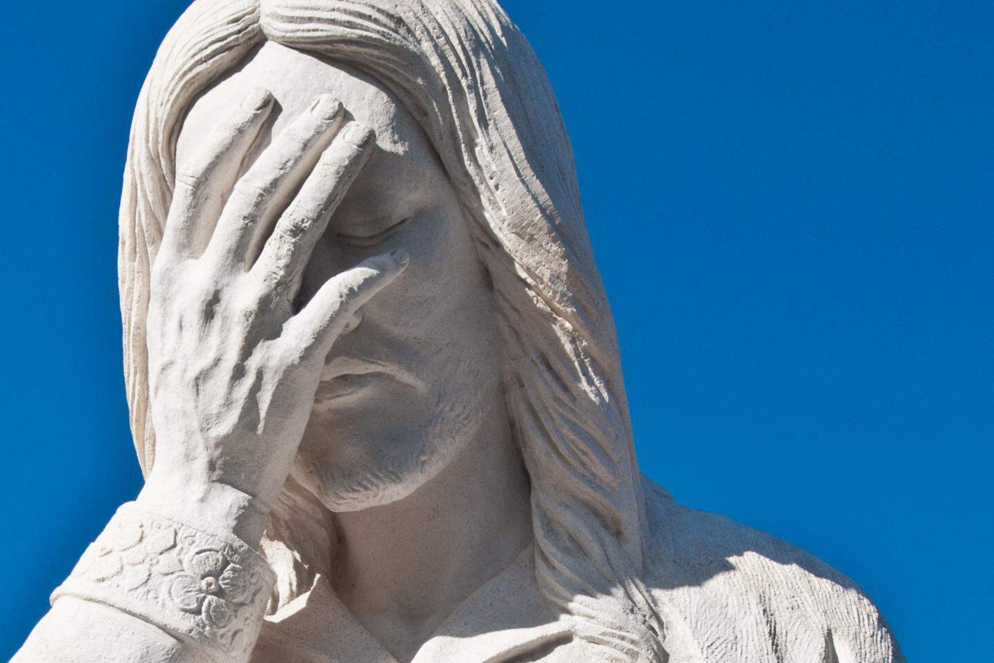 This image shows the face of a statue. The statue is Jesus with a hand up against his face, as if acknowledging something disappointing is happening