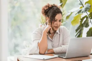 This image shows a woman sitting at a desk with a pen in hand, paper on the desk, and a laptop open in front of her. Her attention is focused on the laptop's screen
